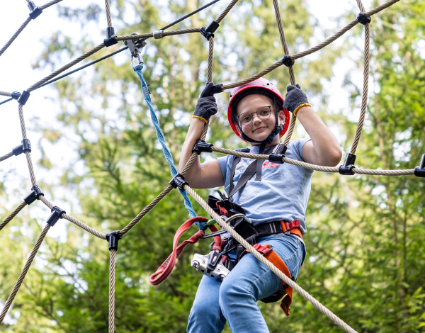Ein Kind mit Kletterausrstung klettert in einem Kletternetz und schaut in die Kamera