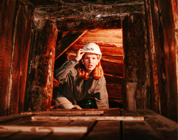 Junge Frau mit Helm schaut staunend durch einen Bergwerksstollen in die Kamera